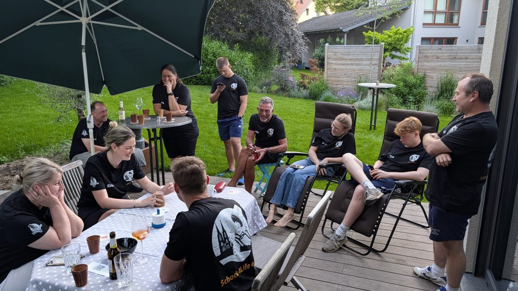 Mehrere Spieler*innen des Schock4Life e.V. sitzen entspannt im Garten und schocken an einem Tisch unter einem Sonnenschirm.