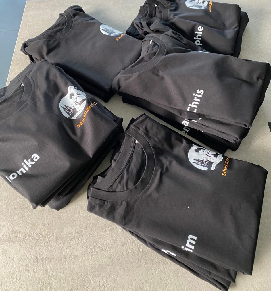 Ein Stapel frisch bedruckter, schwarzer T-Shirts von Schock4Life e.V. mit Vereinslogo und den Namen der Spieler*innen.