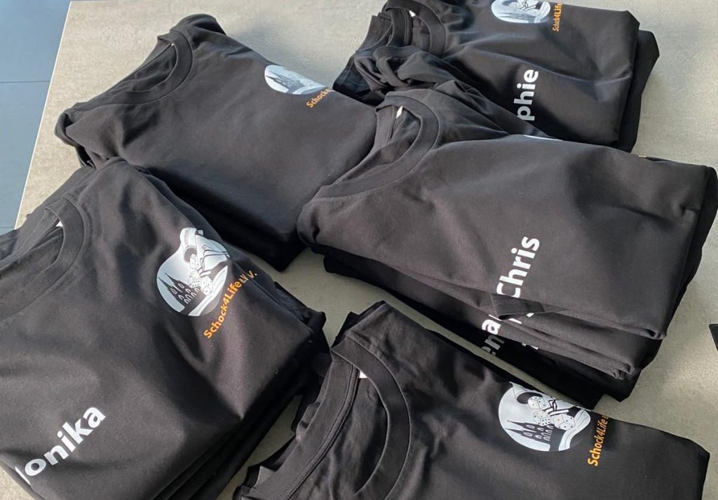 Ein Stapel frisch bedruckter, schwarzer T-Shirts von Schock4Life e.V. mit Vereinslogo und den Namen der Spieler*innen.